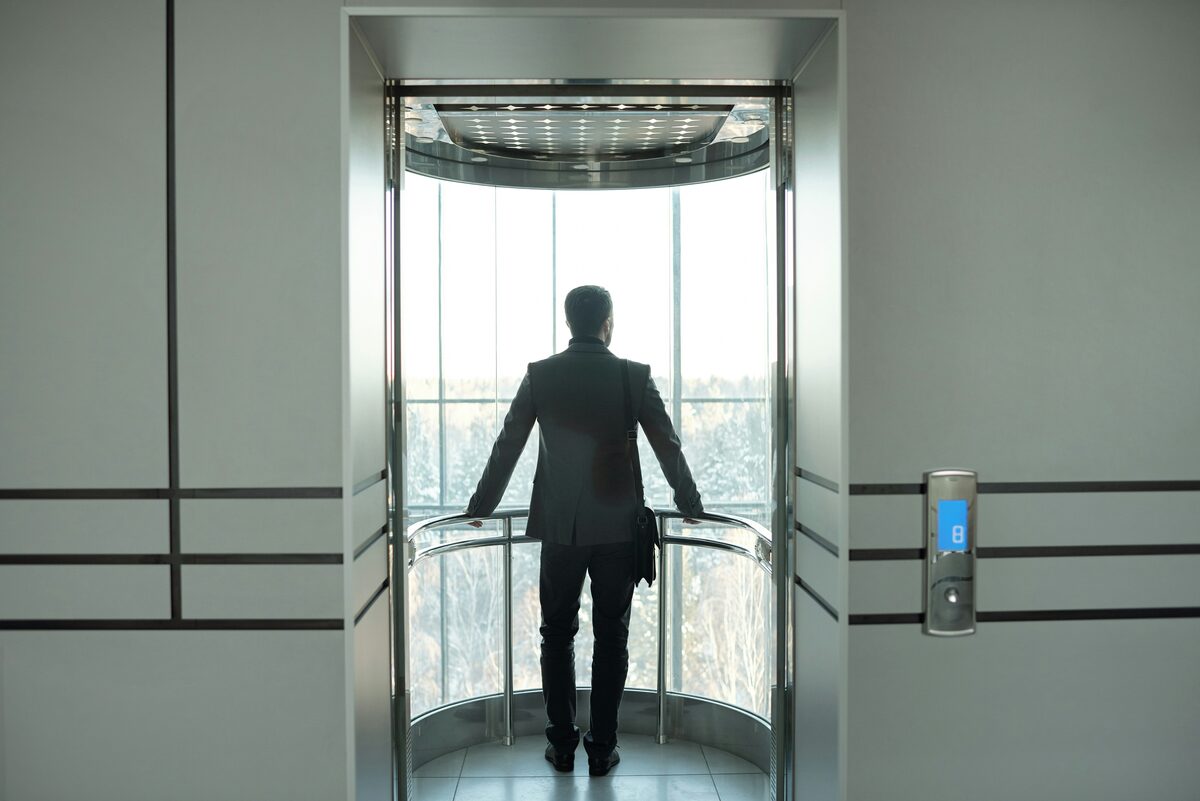 A man with a bag standing in a glass elevator looking out at trees